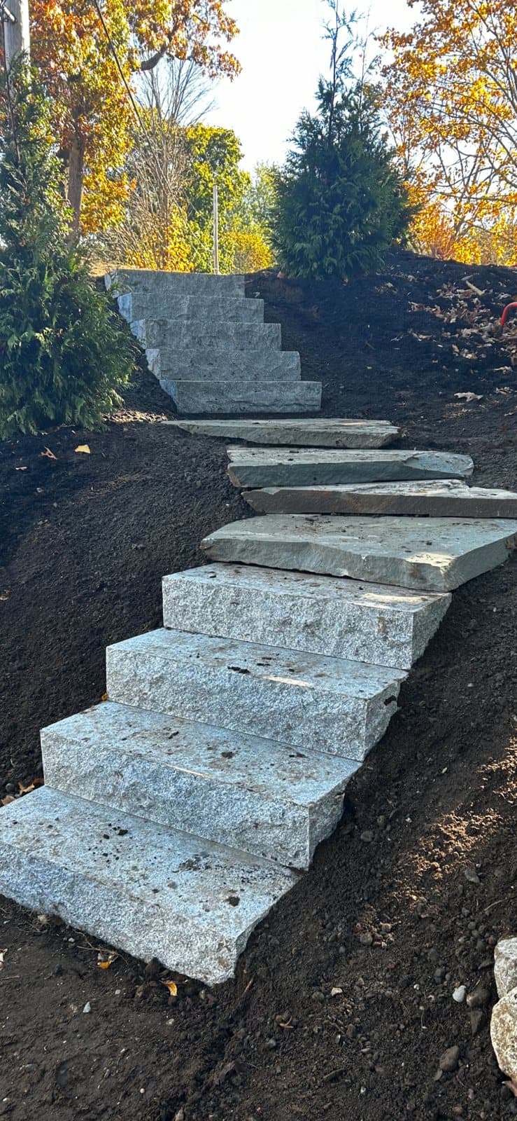 Granite block steps with wide stone treads climbing a freshly loamed slope, with autumn foliage behind.