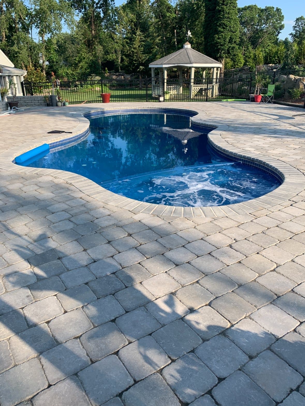 Freeform in-ground pool wrapped in a grey paver patio, with a white gazebo at the far corner.